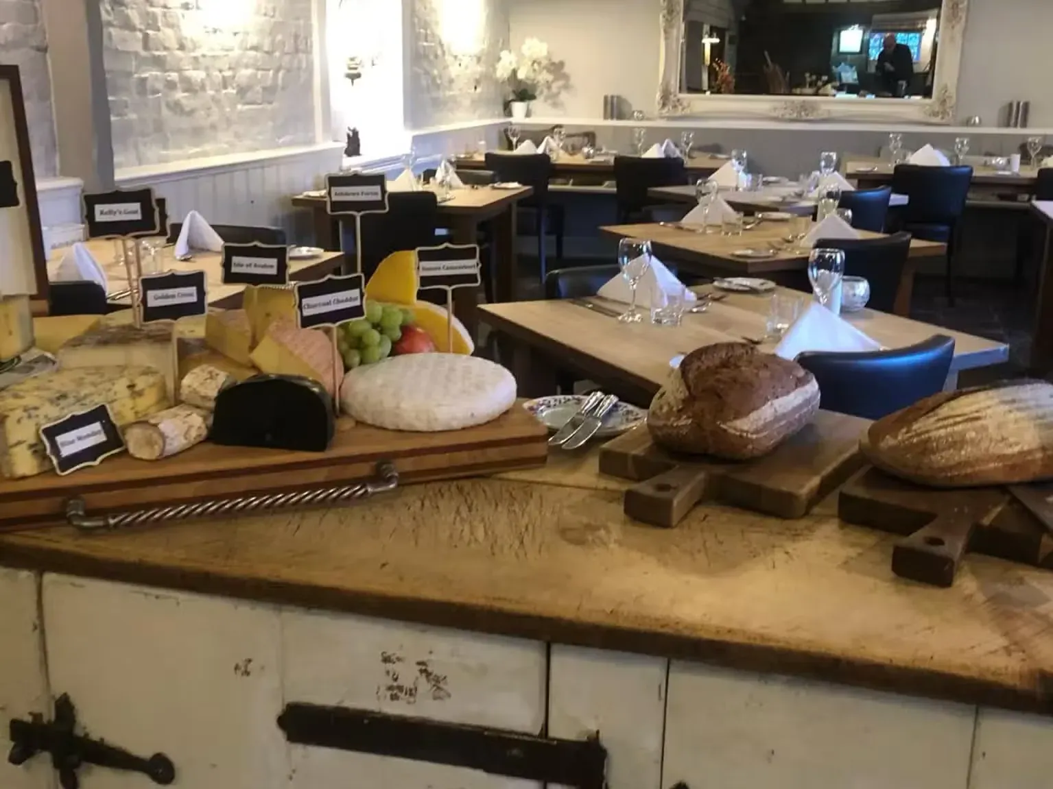 Loaves of bread and a board of cheeses laid out on a wooden counter, with set tables behind.