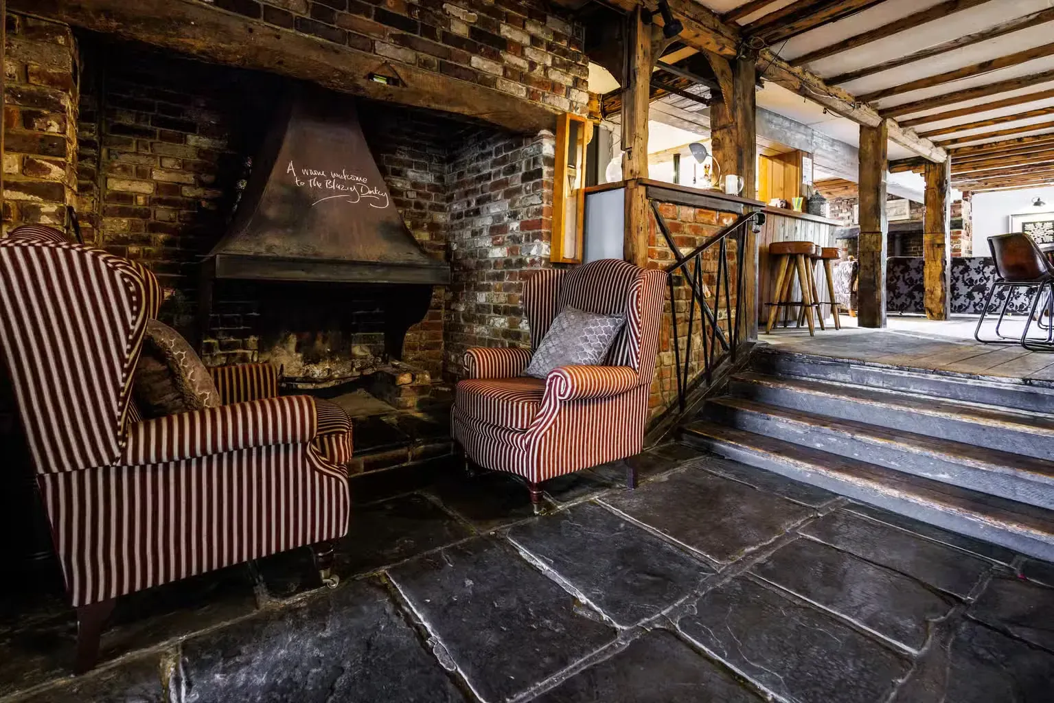 A fireplace corner with two striped wing chairs, an iron rail and worn flagstone steps leading up to the next room.