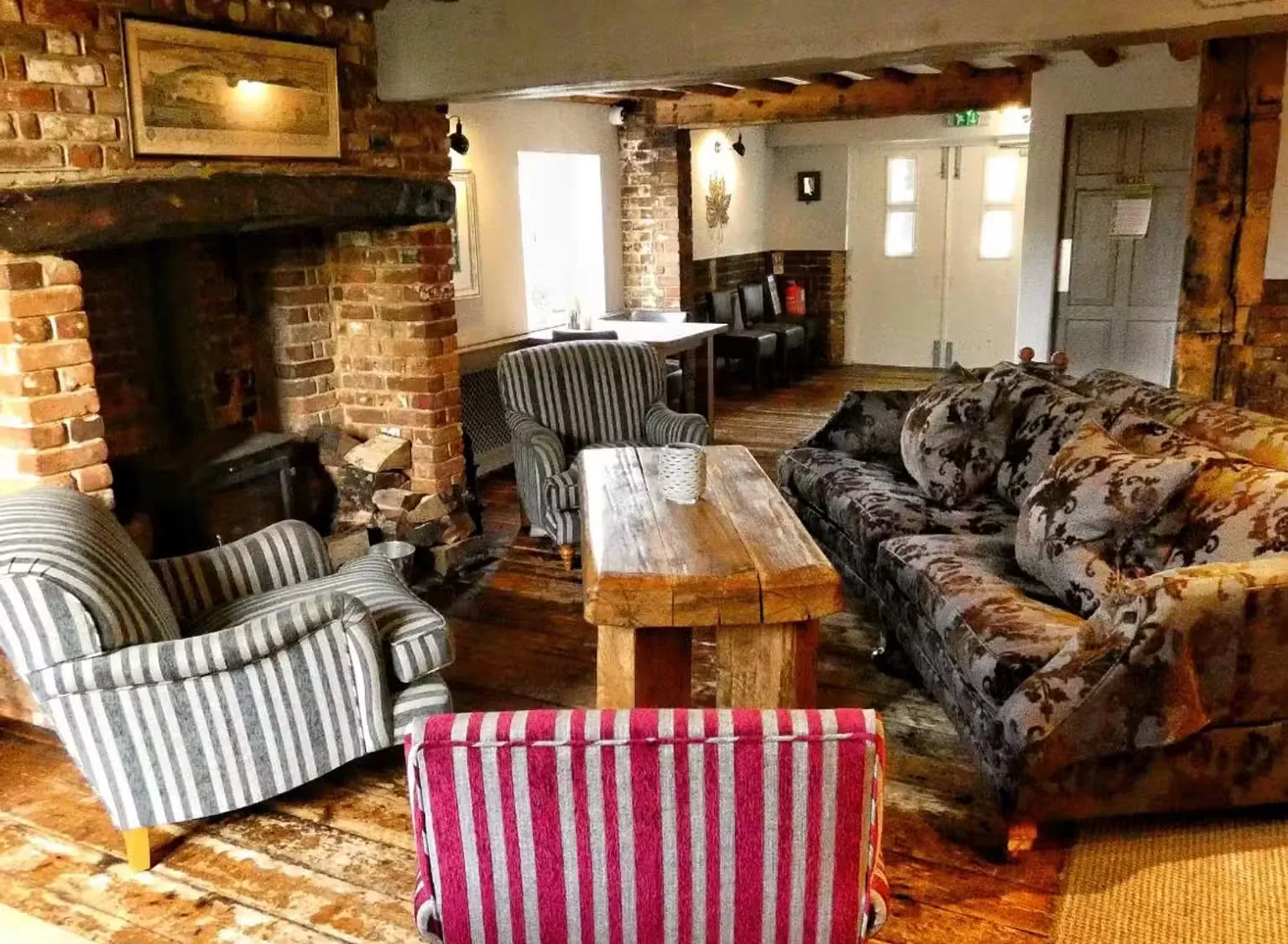 A snug with a brick inglenook fireplace, striped armchairs, a deep patterned sofa and a heavy wooden coffee table.