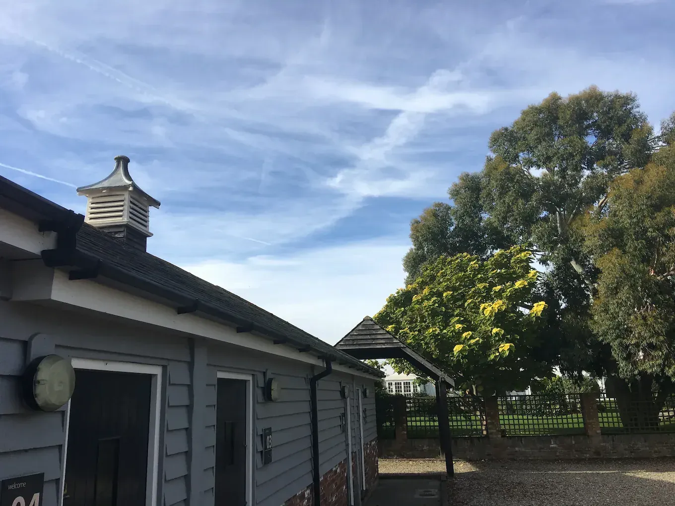 A single-storey weatherboarded stable block with dark doors, standing under mature trees against a blue sky.