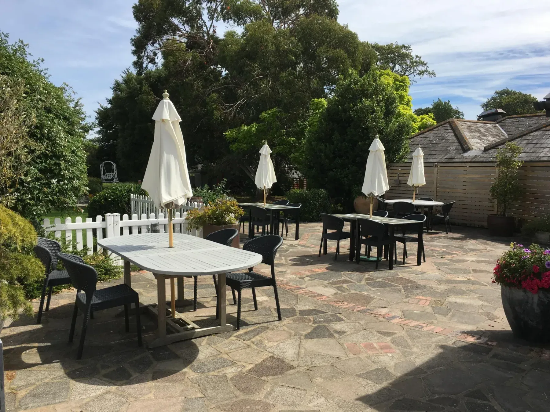 A paved terrace with metal tables and chairs, folded white parasols and pots of flowers, edged by a low picket fence.